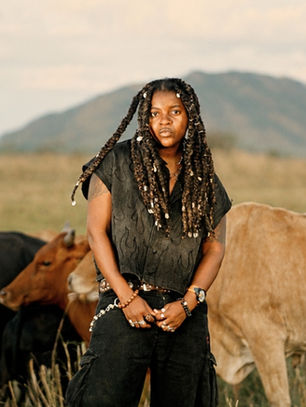 Young woman with long braids stands in a grassy savanna with grazing cattle, mountain in background, calm serious expression.