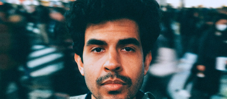 Close-up of a man with a contemplative expression in a busy urban setting; blurred crowd and crosswalk in the background.