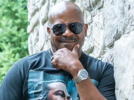 Bald man in sunglasses and black graphic T-shirt poses by a stone wall, smiling thoughtfully with green trees behind.