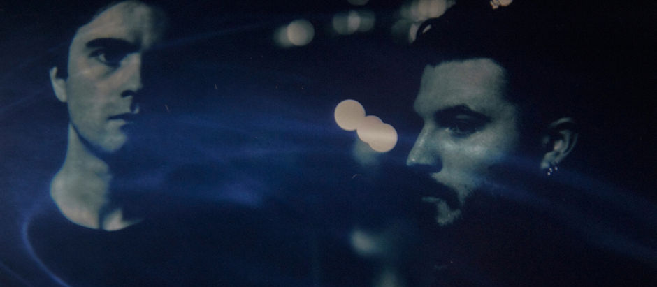 Two men in a dimly lit setting with blue light streaks and blurred city lights in the background, creating a mysterious mood.