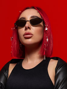 Person with red hair and star earrings wears black sunglasses and leather sleeves against a vibrant red background, exuding confidence.