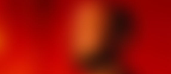 Bald man with beard and earring gazes right against vibrant red background, casting a shadow. Warm lighting creates dramatic mood.