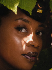 Woman with earrings looks through green leaves, sunlight highlighting her face. She wears dark lipstick and has a confident expression.