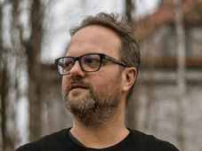 A man with glasses and a beard looks to the side in a tranquil outdoor setting with bare trees and a building in the background. Mood is contemplative.