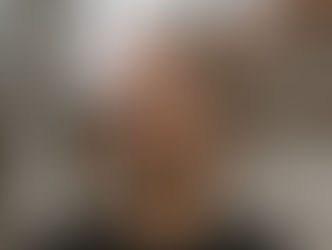 A man with glasses and a beard looks to the side in a tranquil outdoor setting with bare trees and a building in the background. Mood is contemplative.