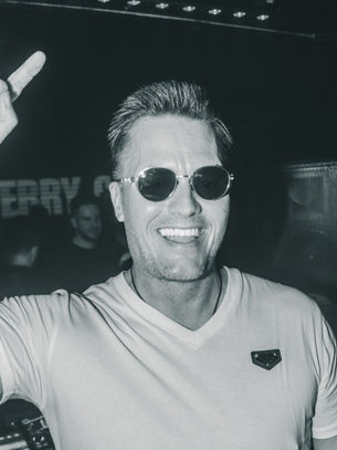 Man in sunglasses and a white shirt throws a rock sign in a dimly lit club. "TERRY" visible on wall. Smiling, energetic vibe.