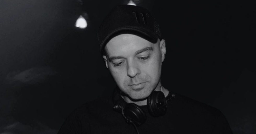 Man in black cap with headphones around his neck, looking down pensively. Dimly lit, dark background with glowing lights above.