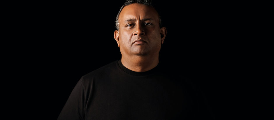 Serious man in a black shirt against a black background, facing forward in dramatic low-key lighting.