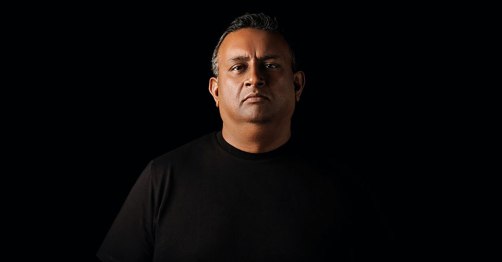 Serious man in a black shirt against a black background, facing forward in dramatic low-key lighting.