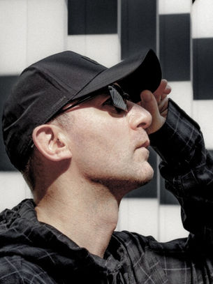 Man in a black cap and sunglasses, looking up. Background has geometric black and white patterns. Casual, contemplative mood.
