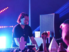 DJ performing at a dimly lit event with colorful lights and mystical decor. A large speaker and decorative objects are visible around him.
