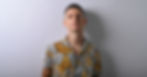Man in a yellow and gray tropical print shirt stands against a plain light gray wall, looking serious.