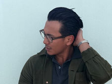 Man in glasses, hand behind head, wearing a green jacket against a pale wall, looking thoughtful.