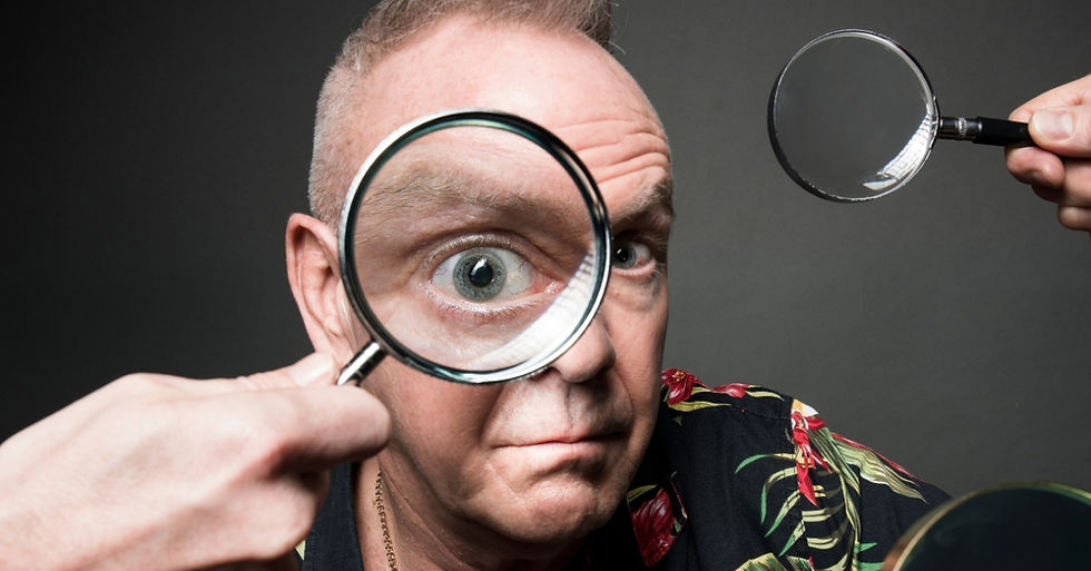 Close-up of a man peering through a magnifying glass while another hovers near his face against a dark background.