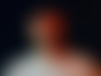 Man in a light shirt and cap stares ahead with a serious expression. Dark background with warm lighting highlighting his face.