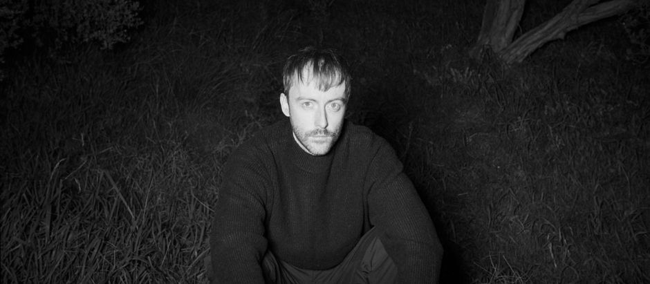 Black-and-white portrait of a serious man crouching in tall grass at night outdoors.