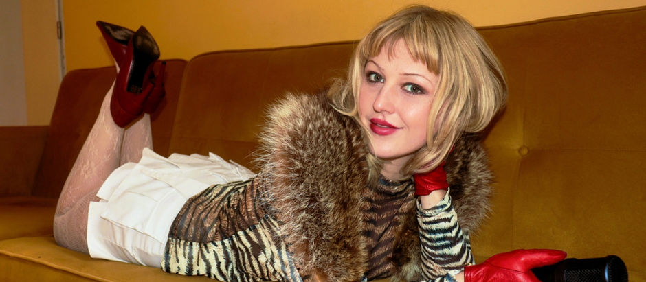 Woman in zebra-pattern top and white skirt, lying on a mustard sofa, holding a microphone. Red shoes and gloves, relaxed expression.