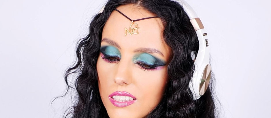 Woman with long black hair wearing white headphones and a gold headpiece. Her eyes are closed, showing vibrant blue and purple eyeshadow.