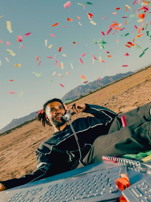 Man lounges in a desert, smiling on a phone as confetti falls, with a mirrored disco ball and laptop in the foreground.