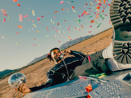 Man lounges in a desert, smiling on a phone as confetti falls, with a mirrored disco ball and laptop in the foreground.