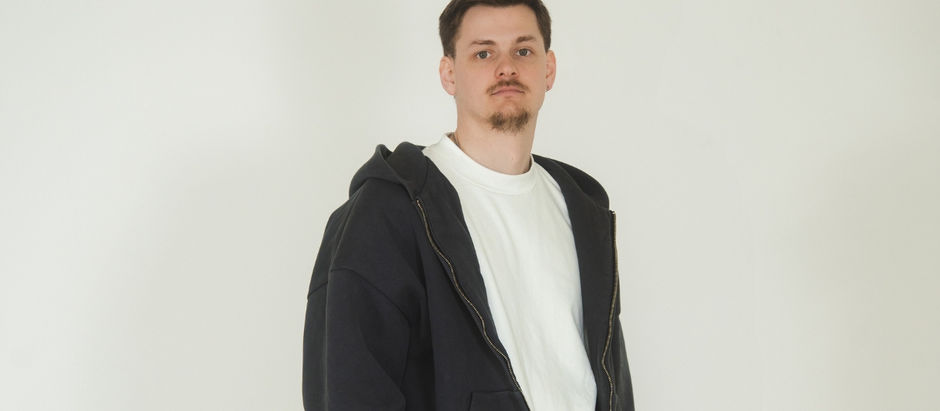 A man in a black hoodie and white shirt stands against a plain white background, looking directly at the camera with a neutral expression.