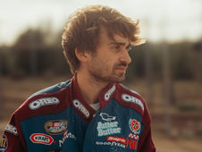 Man in a racing suit adorned with logos like Oreo, standing outdoors with a blurred background, looking contemplatively to the side.