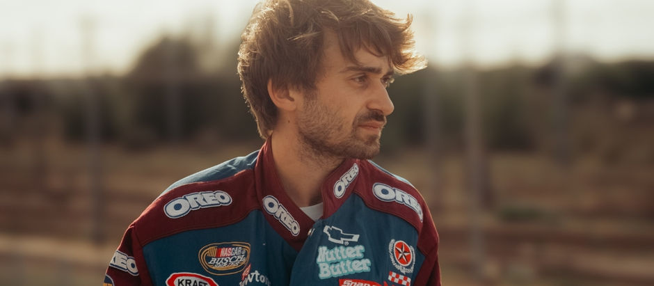 A man wearing a colorful racing suit with Oreo and other logos stands outdoors, looking thoughtful. Blurred background enhances focus on him.