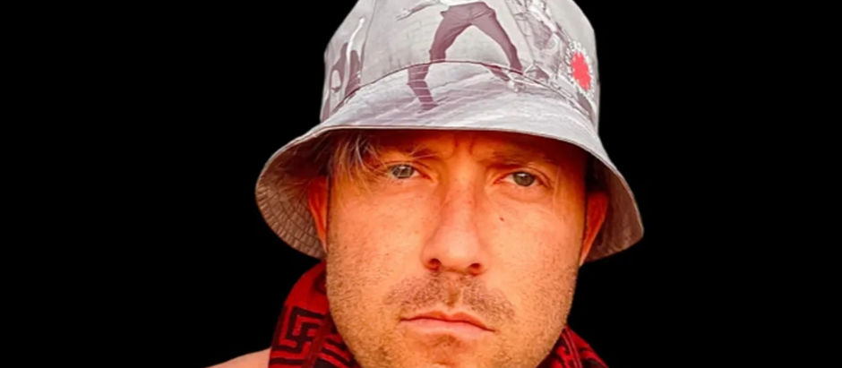 Close-up of a stern man wearing a gray bucket hat and red scarf against a black background.