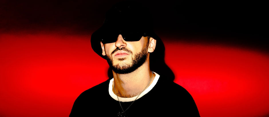 Man in black hat and sunglasses against a vibrant red and black background. He wears a chain, exuding a mysterious, cool vibe.