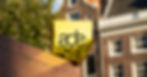 Yellow mirrored sign with black ads logo on a brick wall, with houses and trees softly blurred in the background.