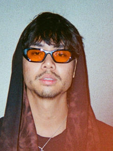Man with orange-tinted glasses and hooded outfit, standing against a plain light gray wall. Red light effect on lower right. Mood is calm.