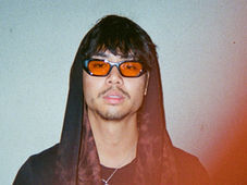 Man with orange-tinted glasses and hooded outfit, standing against a plain light gray wall. Red light effect on lower right. Mood is calm.