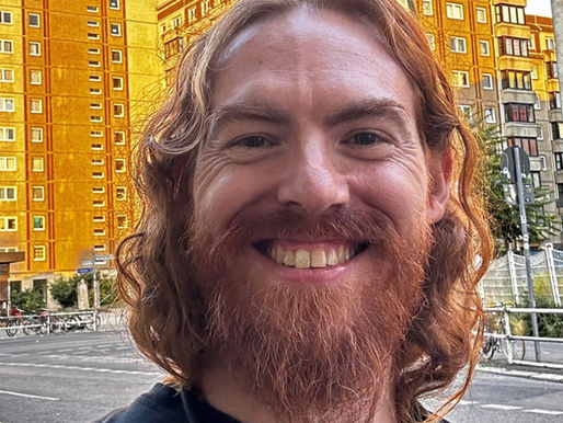 Smiling person with red hair in front of yellow apartment buildings. Urban setting with bikes and graffiti, sign reads "Masser kommt nicht..."