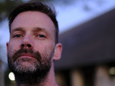 Close-up of a man with a beard, looking serious. Blurred background with a building and trees, sky in soft blue tones.
