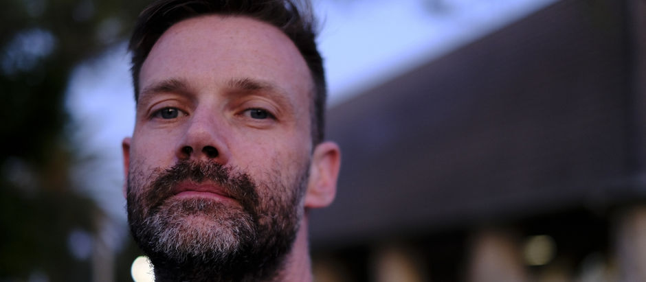 Close-up of a man with a beard, looking serious. Blurred background with a building and trees, sky in soft blue tones.