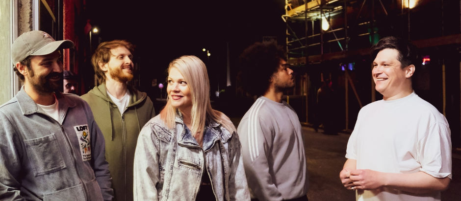 Group of five people chatting and smiling at night on a city street. Dim lighting with scaffolding in the background. Casual attire.