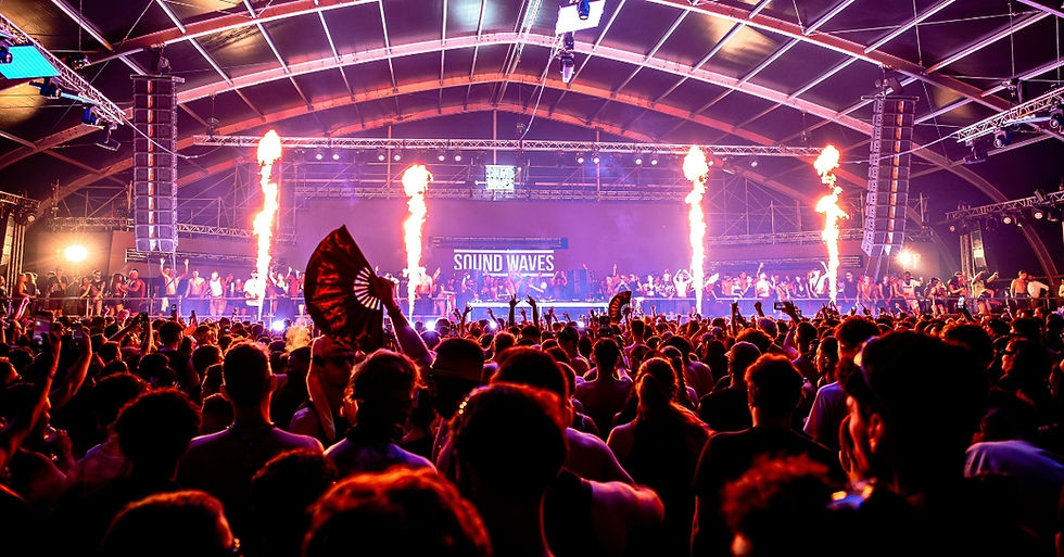 Crowd at a vibrant indoor music festival with pyrotechnic displays. People dance under colorful lights. "Sound Waves" sign visible on stage.