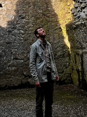 Man stands in a rocky cave ruin, looking up at a shaft of sunlight, with dark stone walls and a quiet, awe-filled mood.