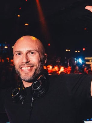DJ with headphones around neck raises hands, smiling in a lively, dimly lit club with colorful lights and cheering crowd in background.