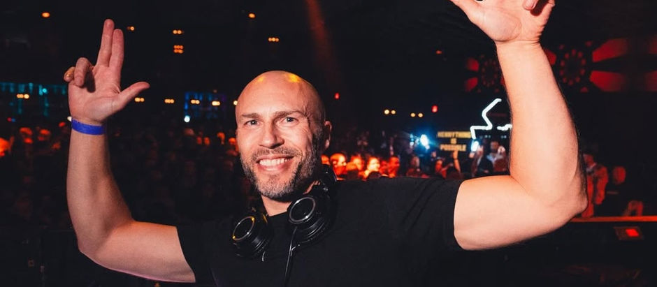 DJ with headphones around neck raises hands, smiling in a lively, dimly lit club with colorful lights and cheering crowd in background.