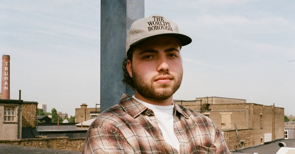 Man in plaid shirt and THE WORLD'S BOROUGH cap on a rooftop, with brick buildings and TRUMAN smokestack behind him.
