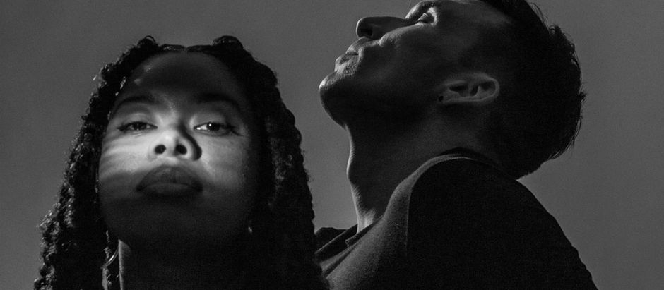 Two people in black and white. One faces forward with light on their face, the other gazes upward. Dramatic lighting and shadows.
