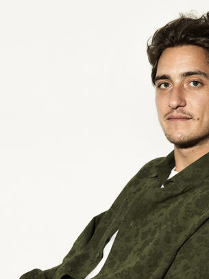 Young man in a green patterned shirt sits against a plain white background, looking forward with a neutral expression. Shadows cast to the right.