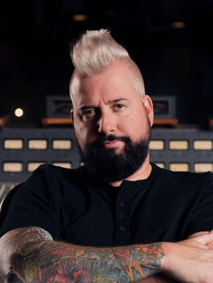 Man with tattoos and styled hair confidently poses in a recording studio, in front of a large sound mixing board and speakers.