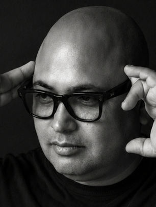 Black-and-white portrait of a bald man adjusting black glasses against a dark background, with a serious expression.