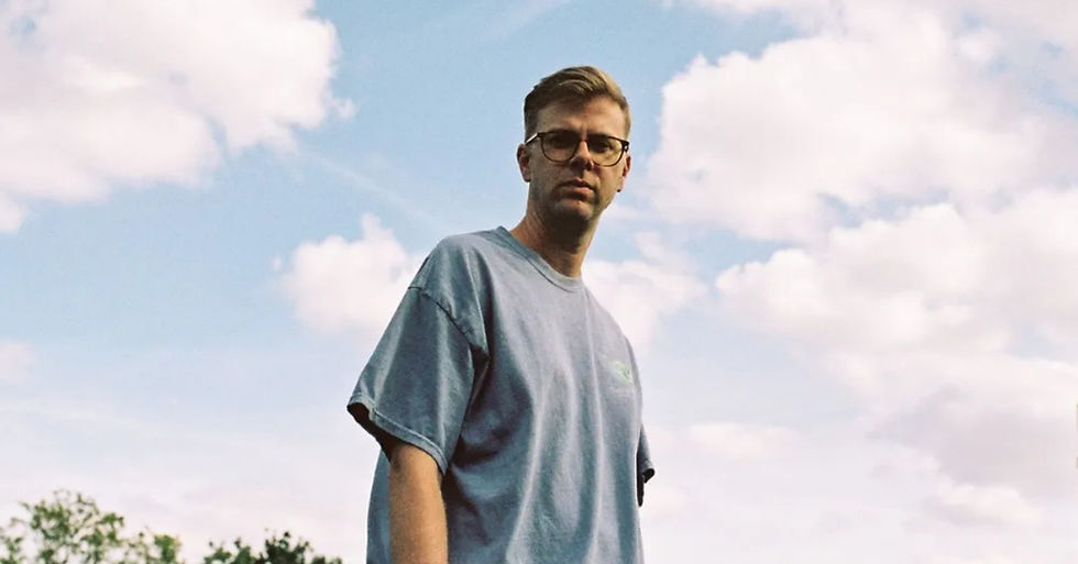 Man in glasses and a gray T-shirt stands against a blue sky with fluffy clouds, looking at the camera.