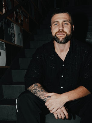 Man with tattoos sits on stairs, surrounded by records on shelves. The setting is dimly lit, creating a contemplative mood.