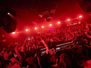 Crowded nightclub under red lights, with a DJ booth, disco ball, and dancing crowd raising hands in a lively scene