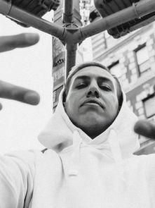 Man in white hoodie makes peace signs, looking confident. Urban background with a tall building and traffic lights. Black-and-white mood.