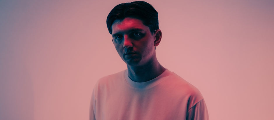 Man in a white shirt stands against a pink and blue gradient background, looking serious. The lighting casts soft shadows on his face.